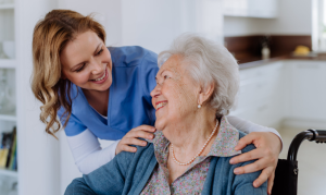 Screenshot 2026-06-17 at 112828 AM - HWK Law Group Caregiver in blue scrubs comforting an elderly woman in a wheelchair with a warm smile in a bright kitchen.